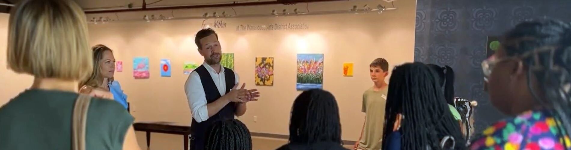 A man speaking to a group of children with a wall lined with art is in the background.