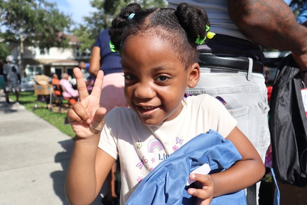 1.jpg Young girl holding a backpack with her left hand and making the peace symbol hand gesture with her right hand.